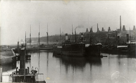 Photograph of the water of the dock at Seaham with a wall of the dock on the left; in the centre is the water on which seven steam boats and one sailing ship can be seen; in the distance, on the right, eight ovens of the bottle works can be seen above the buildings of the town; in the far distance the very indistinct image of th winding gear of a colliery can perhaps be seen