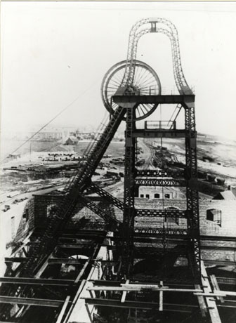 Photograph of the winding gear of a colliery close up ; below the winding gear are struts and round it are walls; in the distance are indistinct buildings and railway tracks; the photograph has been identified as Dawdon Colliery under Construction about 1900