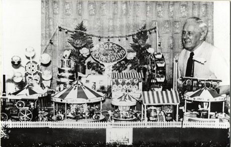 Photograph of an elderly man wearing a shirt and tie standing at the right of the photograph in front of a curtain; in front of the curtain is a miniature fairground of steam engine, three roundabouts, a helter skelter, a big wheel and a sideshow; in the middle is a sign reading Joe's Fairground