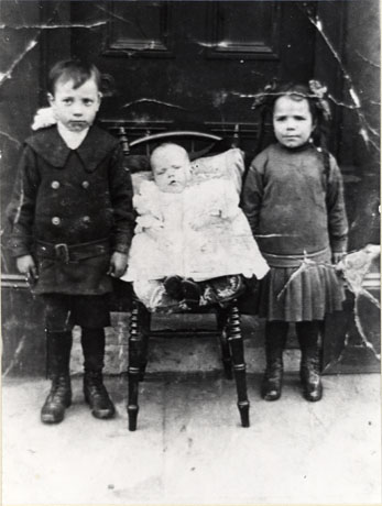 Photograph of a small boy, aged approximately six years, dressed in a double-breasted coat, shorts and boots, on the left of the picture; in the centre is an infant lying on a cushion on a chair, dressed in a robe; on the right of the picture is a small girl of approximately four years dressed in a frock, with a low waist, and boots; they are standing in front of a door; they have been identified as Jim Dawson and sisters, Seaham, 1912