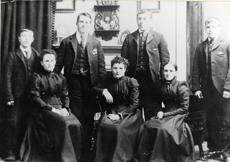 Photograph of four men and three women in the living room of a house; a heavy curtain edged with bobbles is in the left behind two of the people; behind the rest is a chimney piece with a wall bracket above it; the men, who are standing behind the women sitting on chairs, are wearing suits, ties and waistcoats; the women are wearing dresses with long skirts and elaborate bodices; they have been identified as the Dixon-Dawson Family