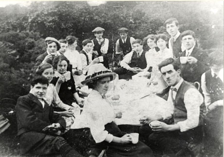 Photograph of seventeen young people sitting round a table covered in a cloth and plates and food; the people are sitting on the ground and behind them are bushes; the photograph has been identified as a Methodist Sunday School outing from Seaham