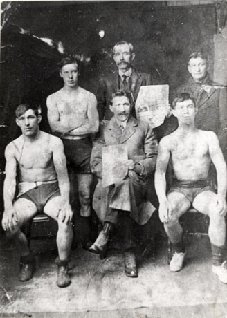 Photograph of three young men wearing shorts, socks and boots only, posed with three men in suits; two of the men are holding sheets of newsprint, which cannot be read; the men have been identified as Local Boxers in Seaham