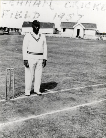 Photograph of a man dressed in cricket whites standing near a set of bails and holding a cricket ball; he has been identified as R. L. Fuller; in the distance two small buildings can be seen; the following is written on the photograph: Seaham Cricket Field, 1957