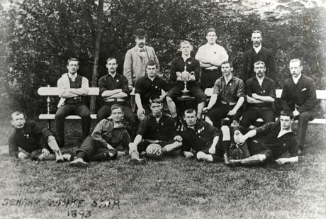Photograph of eleven young men posed on a bench and on the ground in front of trees with four other men; a man in the middle of the group is holding a trophy cup; they have been identified as members of the Seaham White Star Football Team