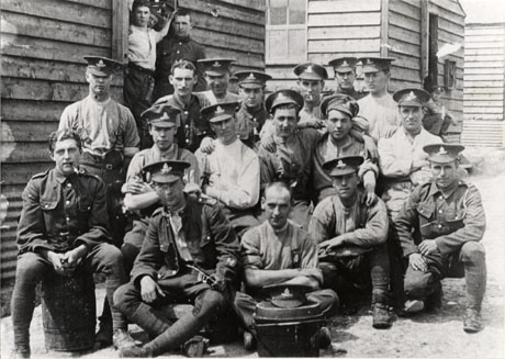 Photograph of a group of seventeen men in soldiers' uniform sitting in an informal group before a row of huts; a number have removed their jackets; three soldiers can be seen in the doorway of the nearest hut and one man is sitting on the step of another hut further away; they have been identified as Seaham Territorials