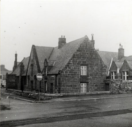 Photograph of the exterior of a stone building showing its end with two windows one above the other, and the side of the building with five windows and a steep gable; two further parts can be seen behind the wing nearest to the camera; the building appears to be lower than the level of the road which runs in front and along its side and front; it has been identified as the National School in Church Street, Seaham