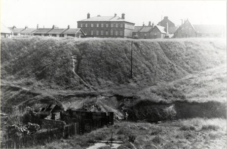 Photograph of a rough grassed slope coming down to an uneven grassed area; on the left of this area are what appear to be allotments; on the crest of the slope are the roof of a low building, a large institutional-looking building, the facade of a row of houses and the end of a church; the photograph has been described as Adam and Eve Gardens, Seaham