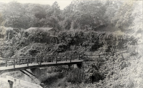 Photograph of a footbridge running from right to left of the picture; behind the bridge is a slope with bushes, and at the top of the slope the roof of a low building; behind that are trees and the roofs of a large house; the bridge has been identified as footbridge at Adam and Eve Gardens, Seaham