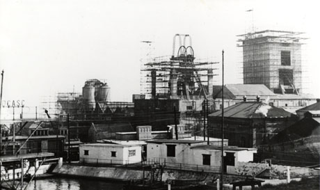 Photograph of the buildings of Dawdon Pit showing scaffolding round the winding gear and two other buildings; in the foreground are low buildings and the surface of water identified as the swimming pool