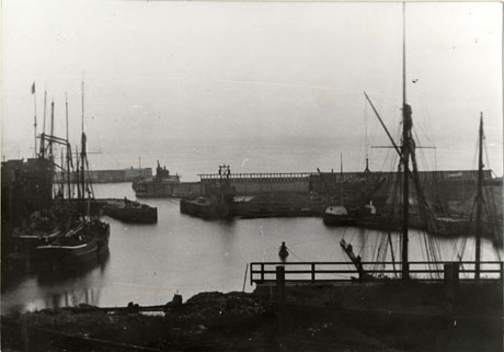 Photograph of a dock looking out to sea, with the walls of the dock running across the middle of the picture; at the side and front of the picture are sailing ships; a boat is being rowed in the dock, which has been identified as Seaham Docks
