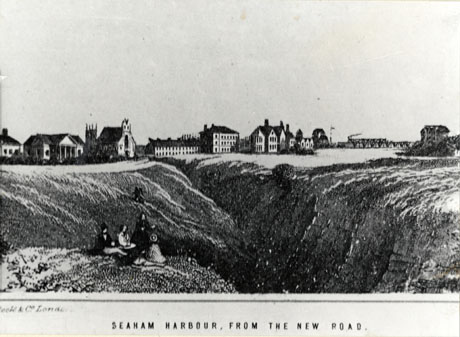 Photograph of an engraving of, in the foreground, a gorge running away from the camera; in the distance are buildings, including a building with a classical facade, a church, a low and a three storey building, a small number of houses and a railway train on a raised track; the picture has a caption : Seaham Harbour, From The New Road and the name Cook and Co., London