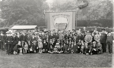 Photograph of approximately a hundred men, women, and children grouped in a field in front of a hedge and large trees; immediately behind them is a miners' banner on which the words Durham Miners' Association can be read and on which a picture of a low building can be seen; a structure with a roof and no sides can be seen behind the group; a policeman is standing on the right of the group; the banner has been identified as being that of Seaham