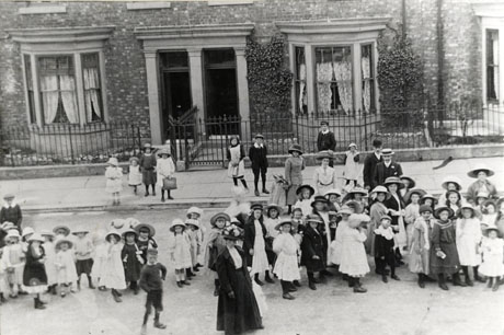 Photograph of a group of approximately fifty one girls dressed in frocks and hats standing in a group the length of a road; ten other children are watching them from the side of the road and from the pavement and six adults are in attendance; behind the figures are the facades of two brick houses with stone surrounds to the windows and doors and railings round the gardens; the photograph is taken from above; the street has been identified as Marlborough Street, Seaham