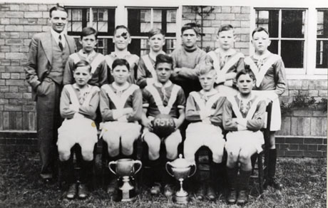 Photograph of eleven boys aged approximately thirteen years in football strip posed with a man outside a brick building; in front of the group are two trophy cups and a boy on the front row is holding a football with the date 1937-38 on it; they have been identified as Dawdon School Football Team