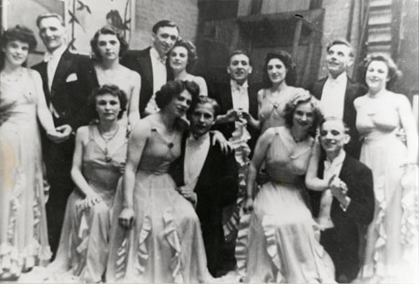 Photograph of eight women in identical evening dresses and six men in white tie and tails; they are seated and standing in front of flats leaning against a wall on a stage; they have been identified as members of the Seaham Amateur Operatic Society