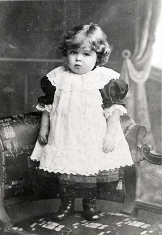 Photograph of a child, aged approximately two years, in pinafore and dress standing on a chaise longue, being steadied by the head rest; he has been identified as Tom Alexander