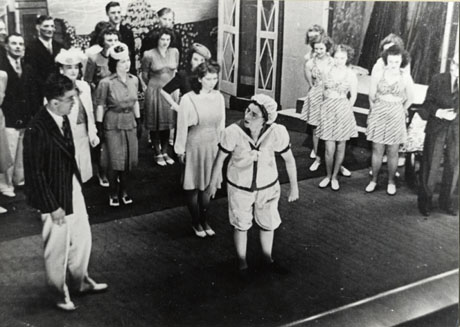 Photograph of the members of cast of a theatrical production on stage in costume; at the front of the stage is a woman in pyjamas, who is talking to a young man in a striped blazer; behind them are six women in the clothes of the nineteen forties and four men in blazers; on the right of the picture are five young women in short skirts and Bikini tops; they have all been identified as members of the Seaham Amateur Operatic Society