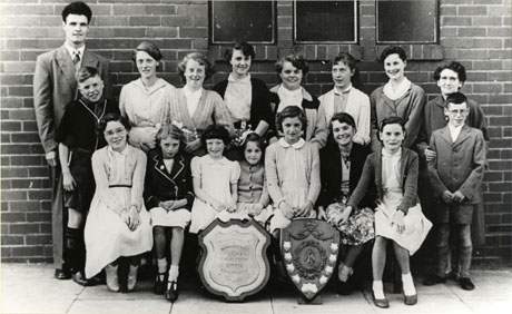 Photograph of a group of twelve girls, aged between six and sixteen years, and two boys aged approximately ten years, posed in front of a brick wall accompanied by a young man and a middle-aged woman; in front of the group are two trophy shields; they have been identified as the winners of the Independent Methodist Challenge Shield