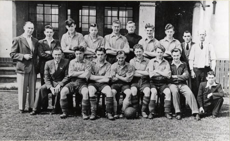 Photograph of a group of eleven young men in football strip, accompanied by four other boys and four men, posed outside the entrance to a building of three steps and doors set back into the building; they have been identified as members of the Dawdon Colliery Football Team