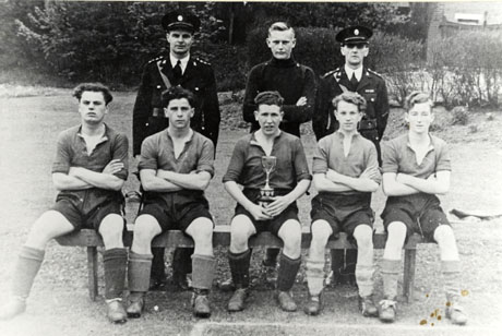 Photograph of five young men in football strip sitting on a bench in the open air with two men in uniform and another man standing behind them; a man on the front row is holding a trophy cup; they have been identified as Dawdon Church Brigade Five-a-Side Football Team