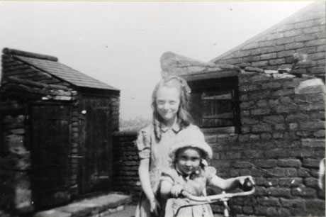 Photograph of a girl aged approximately twelve years standing behind a girl aged approximately six years; the little girl is holding the handlebars of a bicycle and she is wearing a sunhat; behind the girls are a wall with a door for the collection of night soil; further away are two outside privies; they have been identified as the Andrews sisters in Butcher Street, Seaham