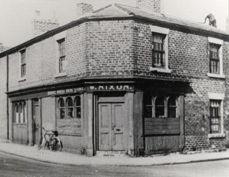 Photograph of the exterior of a shop at the corner of two roads; the facade of the shop is wood, but the rest of the building is brick; three windows and two doorways and the words Bridge House Drug Store W. Nixon can be seen; a bicycle is leaning against the shop; the street has been identified as North Railway Street, Seaham