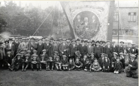 Photograph of a group of approximately sixty men and boys, and two women, in front of the Seaham Lodge banner, displayed in a park with houses behind it; the banner depicts three men whom it is impossible to identify because of the indistinctness of the photograph