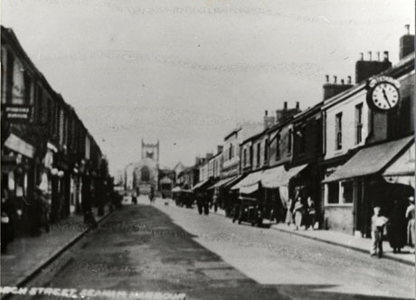 Postcard photograph entitled Church Street, Seaham Harbour, showing the indistinct image of a street receding from the camera; on either side of the street are shops, many with awnings, and a motor car and figures can be seen indistinctly; at the far end of the street is the exterior of a church