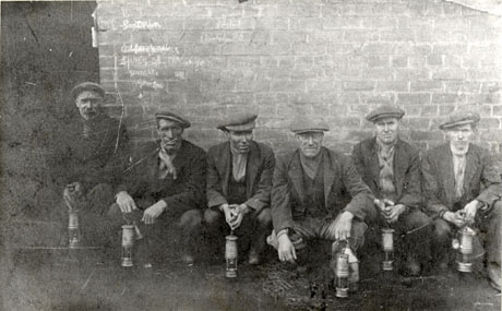 Photograph of six men crouched in front of a brick wall, wearing working clothes of caps, scarves, waistcoats, jackets and trousers; they are all carrying miners' lamps; they have been identified as miners at Seaham