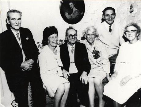 Photograph of three elderly men and three elderly women in front the wall of the living room of a house; patterned wallpaper and the portrait of a young woman are on the wall behind the people; two men are wearing suits and ties and the third is wearing a jumper and tie; the women are wearing dresses and one of the women appears to have a corsage on her dress; they have been identified as the Hoy family of Seaham