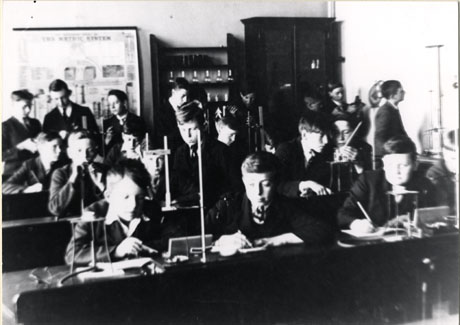 Photograph of approximately eleven boys sitting at laboratory benches with Bunsen Burners and test tubes on them; approximately eight boys are standing at the back of the room looking at items; a table explaining the metric system, an open cupboard containing glass bottles and a closed cupboard are at the back of the room;the boys have been identified as pupils undergoing a Chemistry Lesson at Seaham Modern School