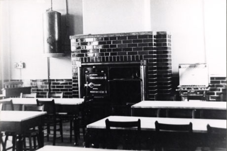 Photograph showing four tables in rows in front of a wall covered half its height in tiles; in the middle of the wall is a chimney breast containing the doors of an oven; at the right hand side of the oven, is a gas stove and on the left hand side, is a water heater mounted on the wall; the room has been identified as Cookery Room at Seaham Modern School