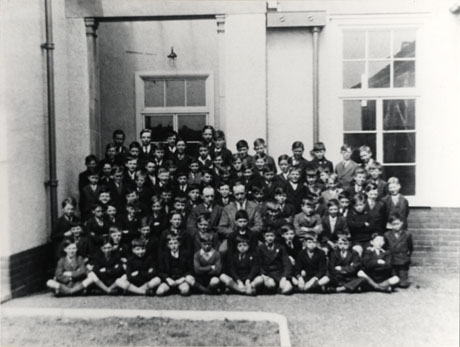 Photograph of a group of approximately eighty boys, aged eleven years, posed against the exterior wall of a building covered in stucco and with large windows; they are accompanied by two men, presumably their teachers; the boys are nearly all wearing jackets and ties; they have been identified as First Year Pupils at Seaham Modern School