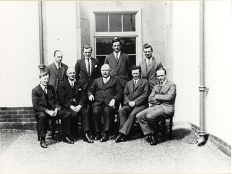Photograph of a group of nine men posed against the wall of building covered in stucco and with large windows; they are all wearing suits and ties; one man is wearing a high collar; they have been identified as Male Staff at Seaham Modern School