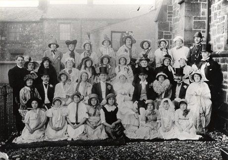 Photograph of a group of twenty nine women and girls, and eight men, in costume, posed with the wall of a church on the right of the picture, and the walls of terraced houses behind them in the distance; railings can be seen on the left of the picture; six of the men are dressed in top hat and tails, one in solar topee and one in a policeman's uniform; the women are dressed long frocks, bonnets and shawls; a clergyman is leaning against the railings; they have been identified as having taken part in a Wesleyan Chapel Concert, Seaham