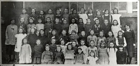 Photograph of forty nine girls, aged approximately ten years, posed in front of a brick building with two women, presumably their teachers; a girl on the front row is holding a notice indicating that the group is part of Dawdon Colliery Girls' School in 1921