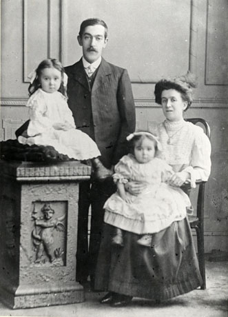 Photograph of a man wearing a suit and tie, standing behind a plinth with a classical design on it; a small girl of approximately two years, wearing a light coloured dress and with ringlets in her hair, is sitting on a cushion on the plinth in front of the man; a woman is sitting on a wooden chair, with arms, to the right of the picture; she is wearing a blouse and long skirt and is holding on her knee a small girl of approximately one year in an elaborate gown; the people have been identified as the Watson Family of Seaham