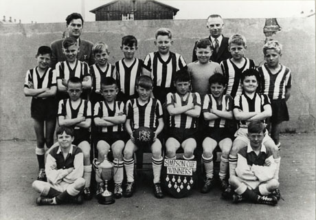 Photograph of twelve boys in striped shirts and shorts, one boy in a jersey, and two boys in long- sleeved shirts in one colour, accompanied by two men, and posed in front of a wall, above which the roof of a wooden building can be seen; a trophy cup and a board on which there are ten medals and the sign Simpson Cup Winners, are on the floor in front of them; a boy on the front row is holding a ball, on which the date, 1962-1963, is written; they have been identified as High Colliery Junior School Football Team, Seaham