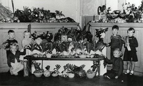 Photograph of two cupboards and a low table in front of the cupboards, covered with fruit and vegetables; three children, aged approximately five years, are either side of the table in front of the cupboards; the photograph has been identified as Harvest Festival High Colliery School, Seaham