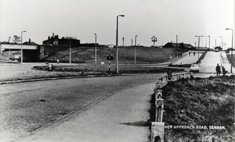 Photograph of a road running from left to right across the photograph; there is a grassed traffic island and grassed strip in the middle of the road, and grassed banks and a railway bridge on the far side of the road; sodium street lights are along the road; two cars can be seen indistinctly on the road; the road is identified on the picture as New Approach Road, Seaham