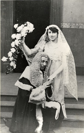 Photograph of a man, dressed in the robes and wig of a judge, kneeling at the bottom of a flight of three stone steps and holding the hand of a woman, dressed as a bride with long veil and bouquet of flowers; they have been identified as Mr. Brownley and Mrs. Elgey, dressed for a performance of Trial By Jury by Gilbert and Sullivan, by the Seaham Amateur Operatic Society