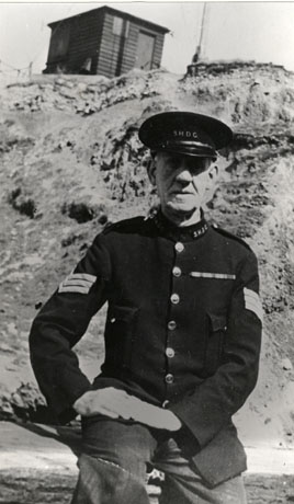 Photograph of a man dressed in a uniform jacket with sergeant's chevrons on the sleeves and the initials S. H. D. C. on the collar; the latter signify, most likely, the Seaham Harbour Dock Company; he is wearing a peaked cap bearing the same initials; behind him is cliff at the top of which is a small wooden hut; he has been identified as Mr. Gatenby, Seaham Docks