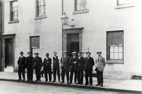 Photograph of ten men standing in a line on the edge of the pavement in front of a the facade of a building covered in stucco with six long sash windows and two entrances, above one of which there is a lantern, and beside which there is a plaque with the inscription: Seaham Harbour and District Social Club; the club has been identified as being in North Terrace, Seaham