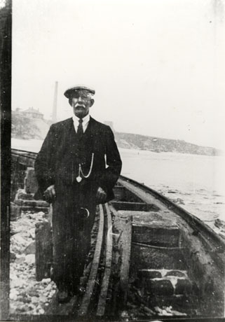 Photograph of a man with a white moustache wearing a suit, tie and cap, standing next to a wooden rail curving to the left behind him with the sea beyond it; in the distance are hills running down to the sea and a tall chimney on top of the hills; he has been identified as William Cole of Seaham, showing the blockyard railway