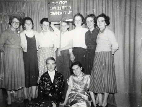 Photograph of seven girls and women standing in front of a curtain arm in arm; in front of them two girls dressed in oriental dresses are sitting on the ground; one of the girls standing up is holding a placard with the words Junior Christian Endeavour on it; they have been identified as members of a Junior Christian Endeavour Concert Party