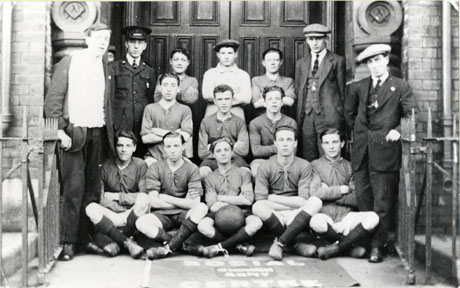 Photograph of eleven members of a football team posed in the doorway of a building with Masonic symbols on its portals, identified in seah0172 as the Candlish Hall; four men, including one in uniform, are standing with the members of the team; in front of the group on the floor is a mat which appears to have the following words on it: Social Centre [Salvation] Army; the people have been identified as members of the Church Army Football Club