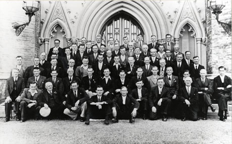 Photograph of approximately sixty five men of all ages posed in front of the exterior of the door, and part of the wall, of a Gothic church building; they are all dressed in suits, or trousers and jackets; they have been identified as Seaham and District Miners