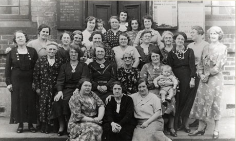 Photograph of twenty women of all ages dressed in frocks posed outside a brick building with a notice board announcing church services on it; three of the women are holding infants; they have been identified as celebrating the twenty- first anniversary of the Womens' Auxiliary Independent Methodist Church, Seaham