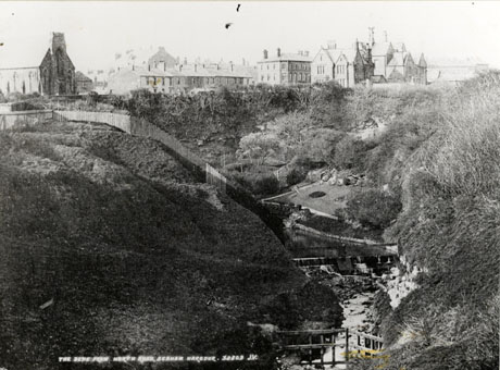 Postcard photograph entitled The Dene From North Road, Seaham Harbour. 32809 J.V., showing a gorge with steep wooded sides and a stream at the bottom; a fence is running down the slope on the left of the picture; in the distance on the top of the cliff, a church, rows of terraced houses, and a large imposing house can be seen